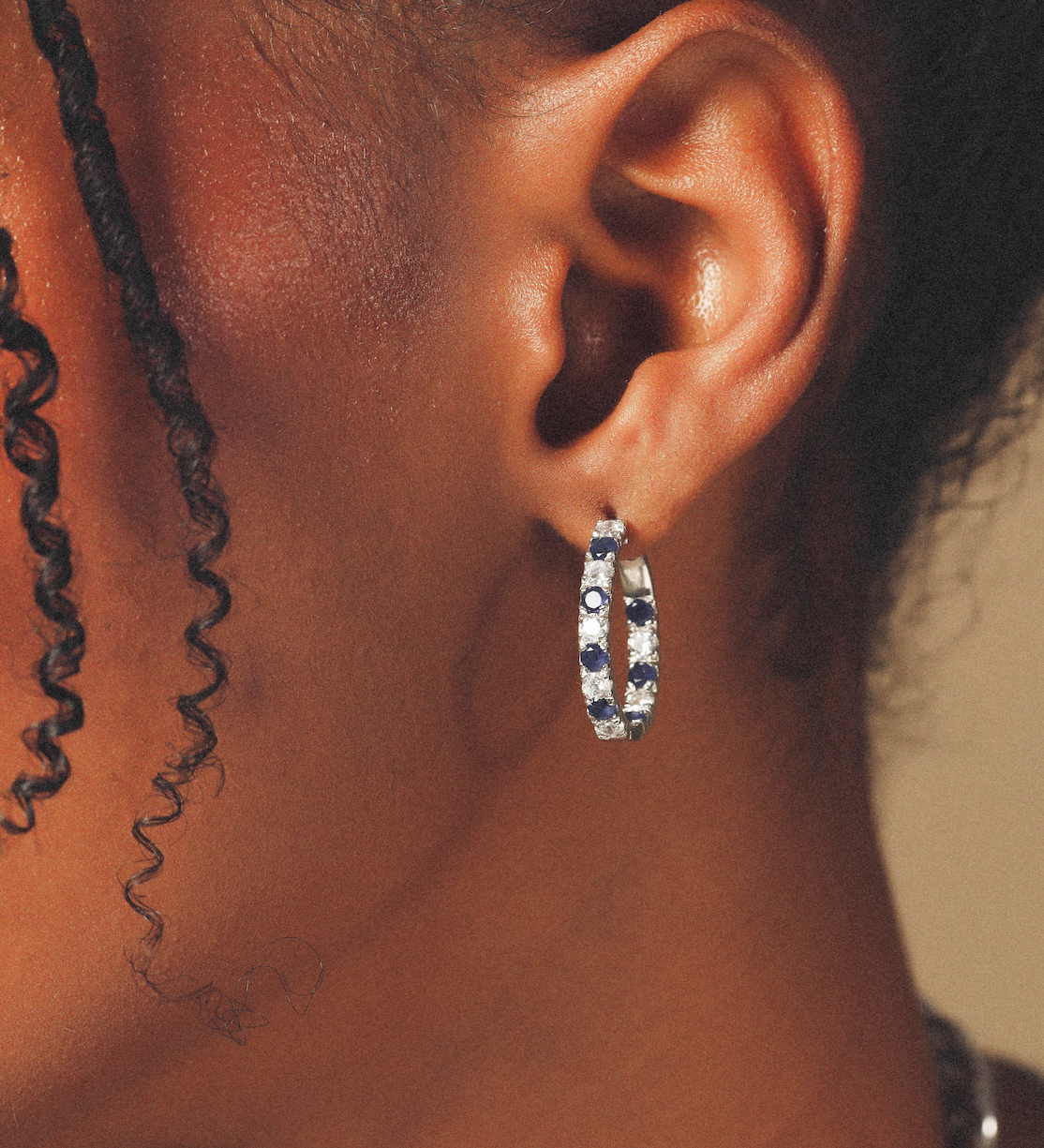 Close-up of an ear wearing a silver hoop earring with blue stones on a neutral background