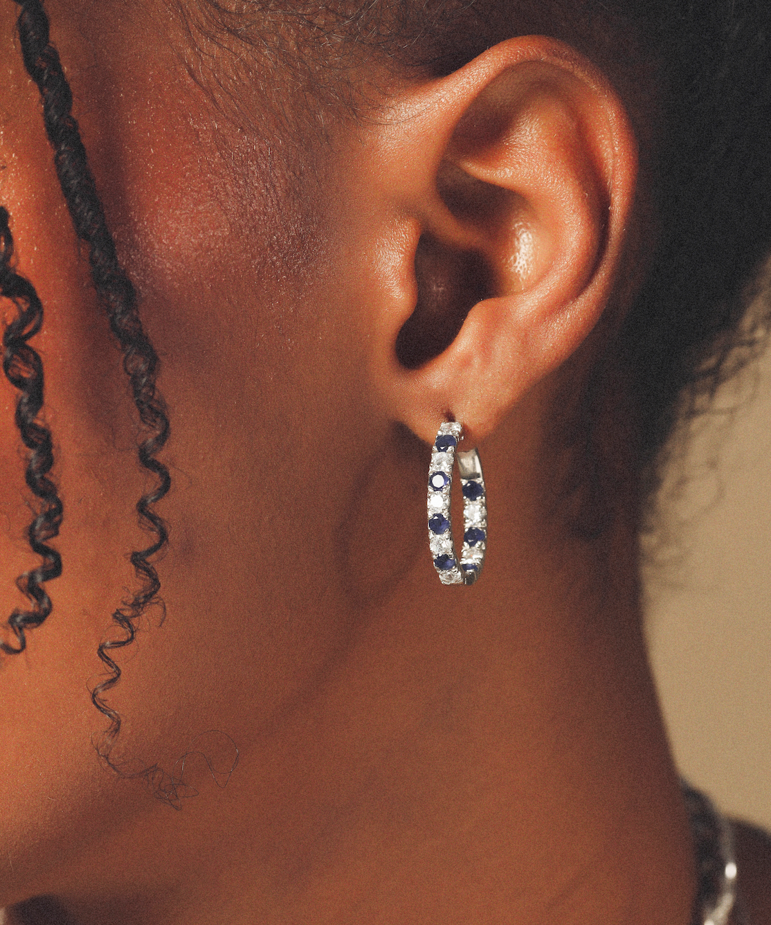Close-up of an ear wearing a silver hoop earring with blue stones on a neutral background