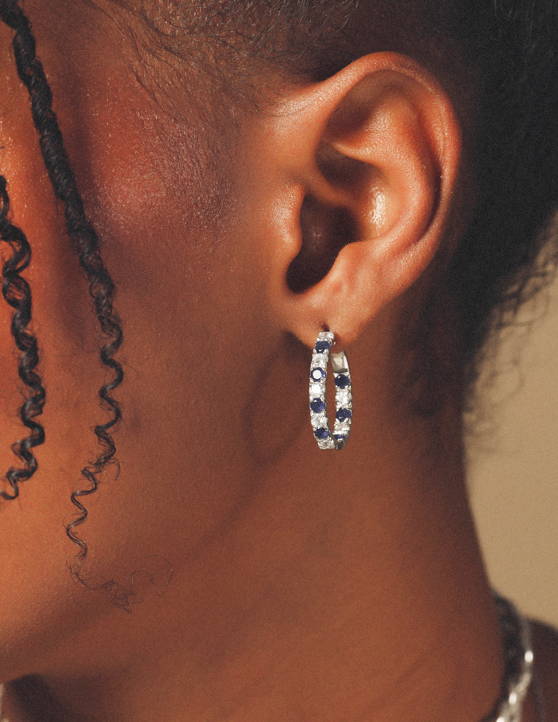 Close-up of an ear wearing a silver hoop earring with blue stones on a neutral background